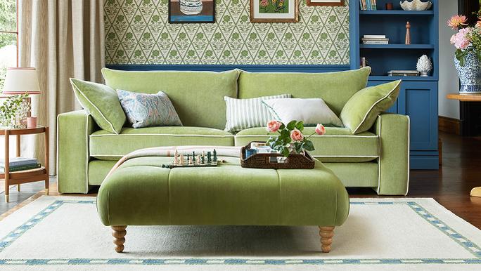 A green sofa with matching footstool in front of a patterned wallpaper