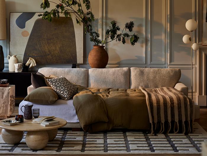 Cream sofa bed with brown bedding on a monochrome patterned rug with a round marble coffee table next to a panelled wall