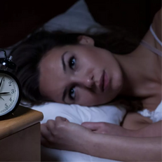 Person laying in bed staring at an alarm clock reading 3:05am