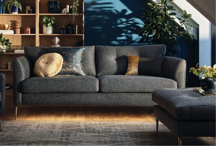 A modern living room featuring a dark grey sofa with decorative gold and blue pillows, matching footstool, and a wooden bookshelf with plants 