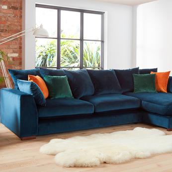Velvet blue corner sofa with orange and green velvet scatter cushions next to an interior brick wall and fluffy white rug