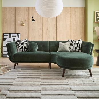 Olive green chaise end sofa with monochrome scatters on a patterned rug with wooden panelling on the wall behind