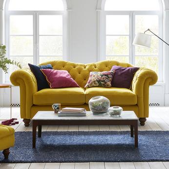 Yellow chesterfield sofa with colourful scattered some with flowers on next to a bright large window sat upon a blue rug