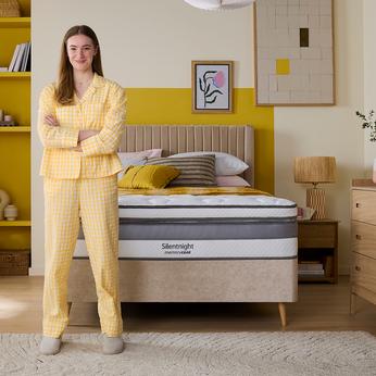 A women stood in yellow pajamas in front of a beige divan bed with yellow walls with a Silentnight mattress 
