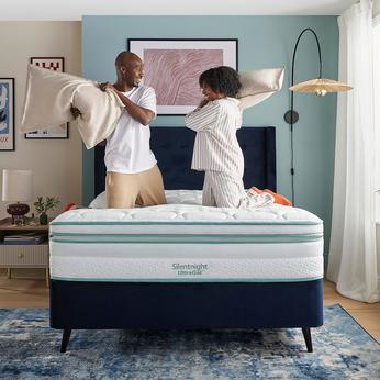 A young couple holding two pillows and standing on a black divan bed having a pillow fight on a silentnight mattress