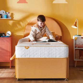 A young boy sat on a orange single bed with a circular headboard, with orange walls with clouds on a kids Silentnight mattress