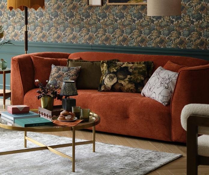 70s style curved sofa in orange with button detailing next to a green floral wall with gold and floral accessories and scatters