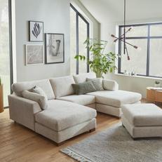 Light grey u shaped fabric corner sofa with square footstool in a bright living room with large windows 