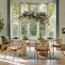 Oak dining table and chairs with spindles next to a large glass window and a hanging floral plant