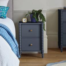 Navy blue bedside table with bronze handles with a plant and next to white and blue bedding