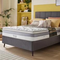 Light grey divan bed next to a yellow wall with a Silentnight mattress with yellow and orange cushions