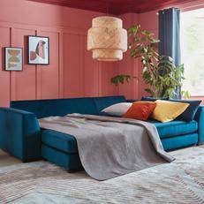 Blue velvet corner fold out sofa bed with bright coloured velvet scatter cushions in orange and yellow next to a pink wall