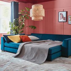 Blue corner sofa bed next to a bright pink wall with orange and yellow scatter cushions and grey bedding