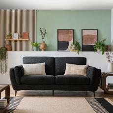 3 seater black sofa with black metal legs next to a pale green wall with white scatter cushions and a beige rug