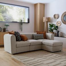 Beige modular corner sofa with orange and monochrome scatter cushions on a wooden floor with a large window