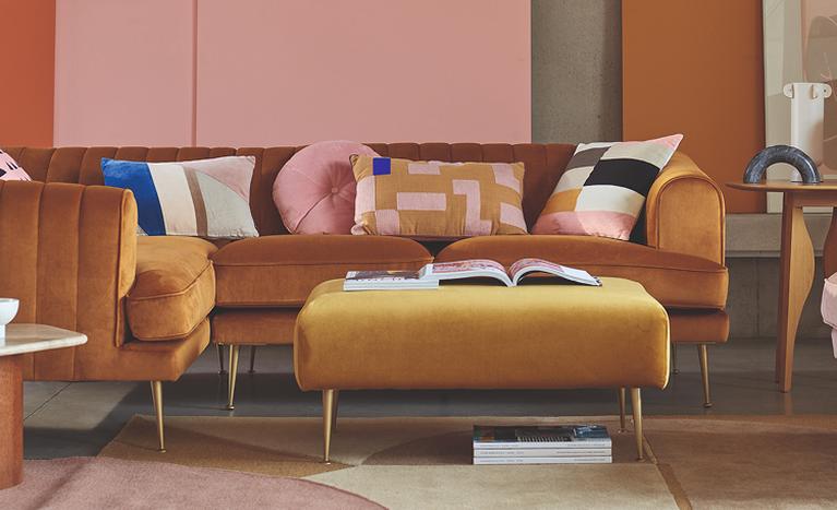Yellow velvet corner sofa with gold metal legs next to a pink wall with matching velvet footstool