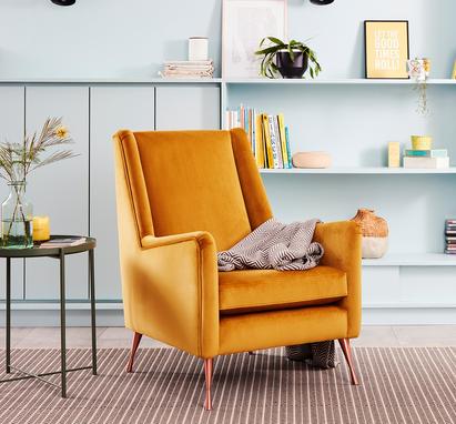 Yellow velvet accent chair with gold metal legs on a stripy rug next to a pale blue wall