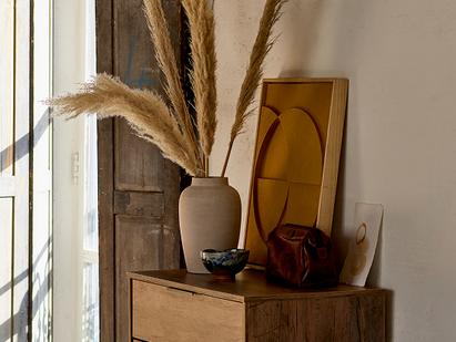 Cream vase with dried pampas grass, art and a wooden bowl sat on top of a dark wood chest of drawers