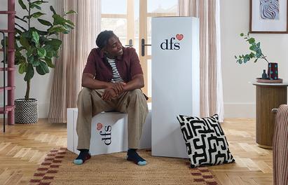 A young man in a pink shirt and white trousers sat admiring his small living with his new sofa in boxes