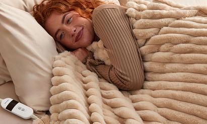 A women with red curly hair curled up into a fluffy beige heated blanket