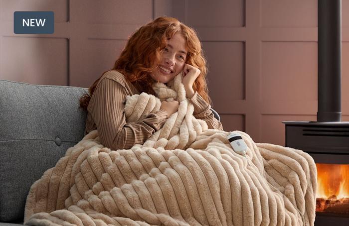 A young women curled up on a grey sofa next to a log burner in a fluffy cream faux fur heated throw