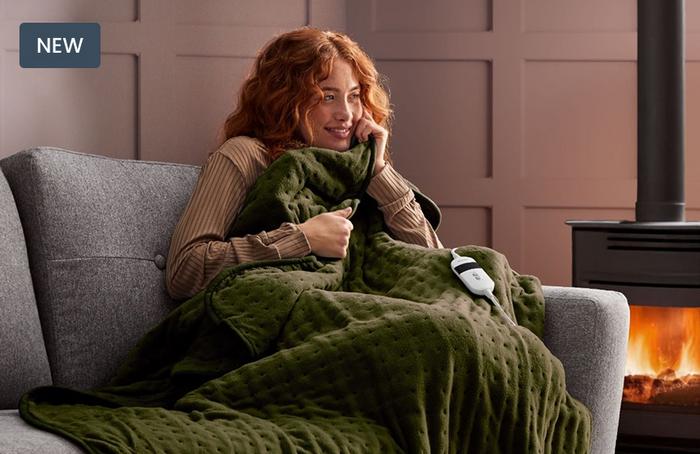 A young women curled up on a grey sofa next to a log burner in a fluffy olive green heated throw