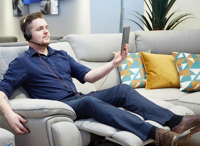 A shop worker reclining on a grey sofa with headphones on and a tablet as he talks to a customer from the shop
