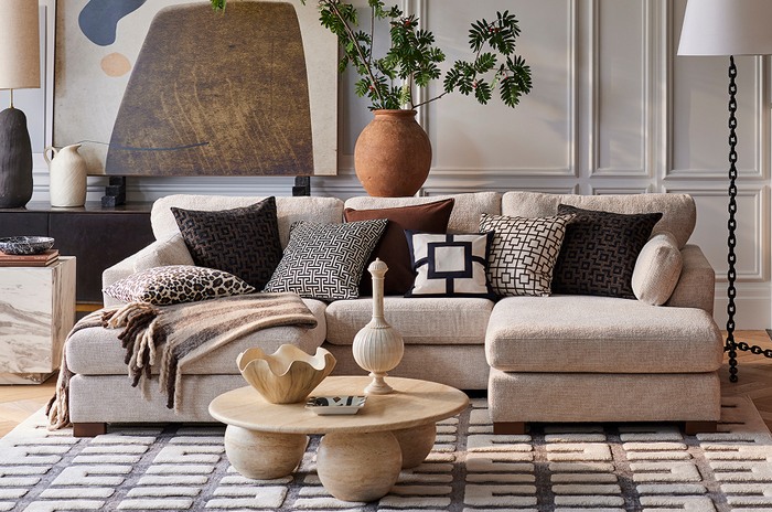 Beige u shaped corner sofa with a mixture of brown, monochrome patterned cushions on a monochrome rug and a round coffee table