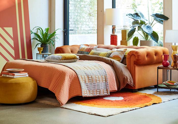 Orange chesterfield sofa bed with a round orange footstool and colourful bedding sat upon a bright orange rug