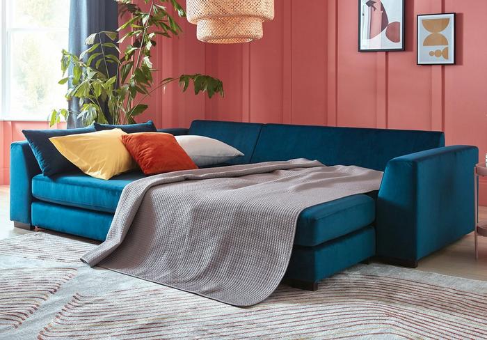 Blue velvet sofa bed next to a bright pink paneled wall with orange and yellow scatter cushions