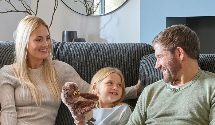 Male and female smiling sat on a sofa looking at a young girl also smiling whilst holding a teddy bear