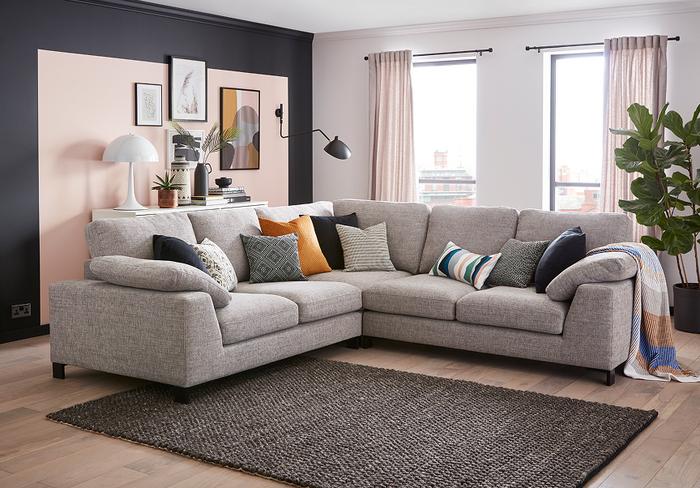 Light grey corner sofa with a variety of monochrome and orange scatter cushions in a bright living space with a pink and grey wall and monochrome rug