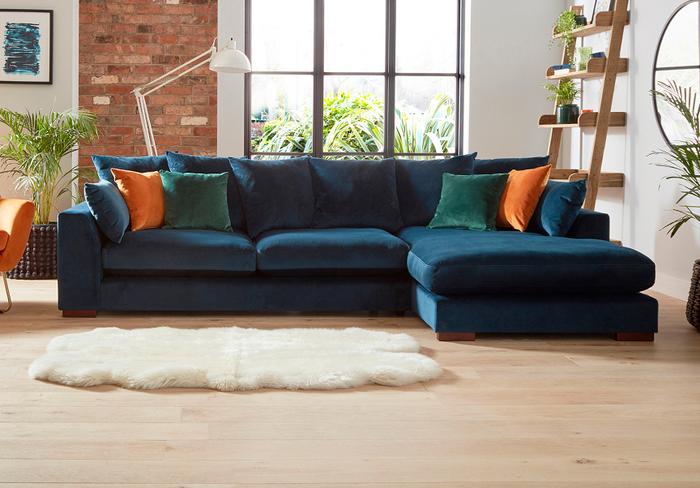 Navy blue velvet corner sofa with orange and green velvet scatter cushions next to a white fluffy rug