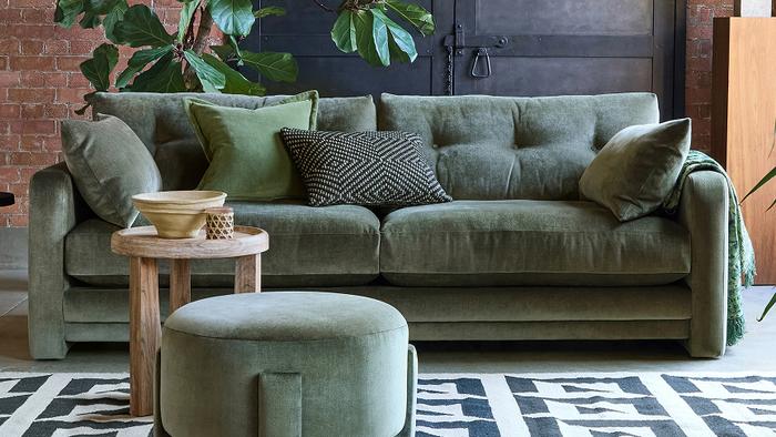 Dark green fabric sofa with a round footstool next to a light wood side table on a patterned monochrome rug
