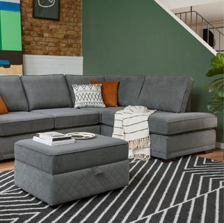 A grey corner sofa with orange and white cushions, paired with a matching ottoman, set on a geometric black and white rug.
