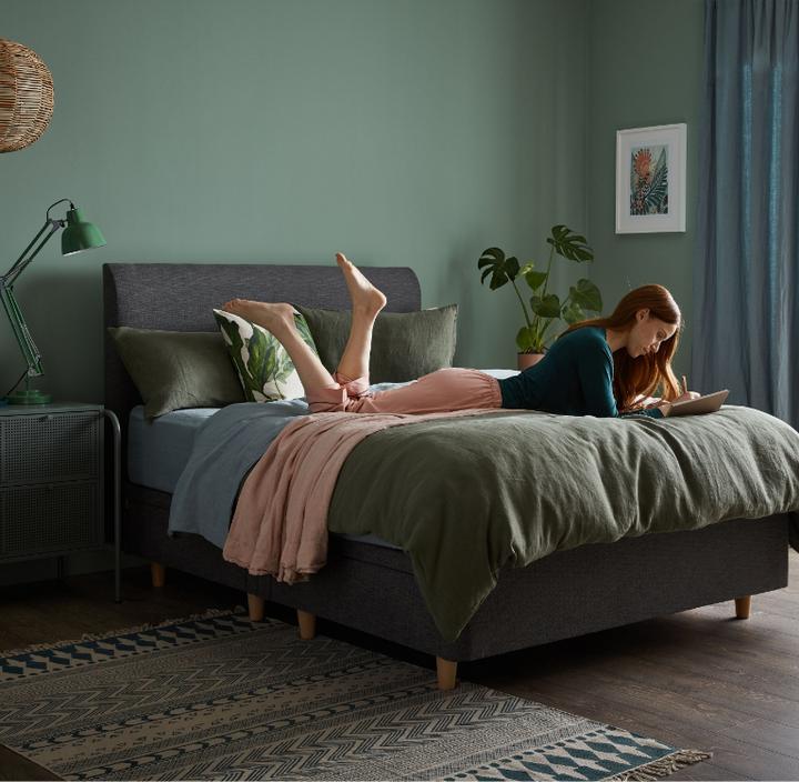Young women laid across a grey divan bed with dark green bedding writing in a notepad