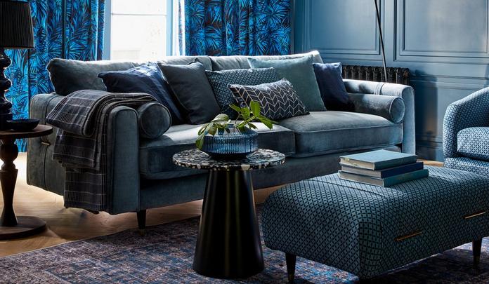 Blue chenille sofa inside a dark blue room with patterned curtains and footstool