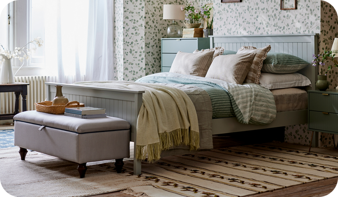 Pale green wooden slat bed with patterned footstool against a patterned wallpaper wall and black and beige rug