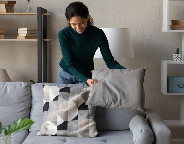 a young women smiling as she plumps and turns cushions on a grey sofa