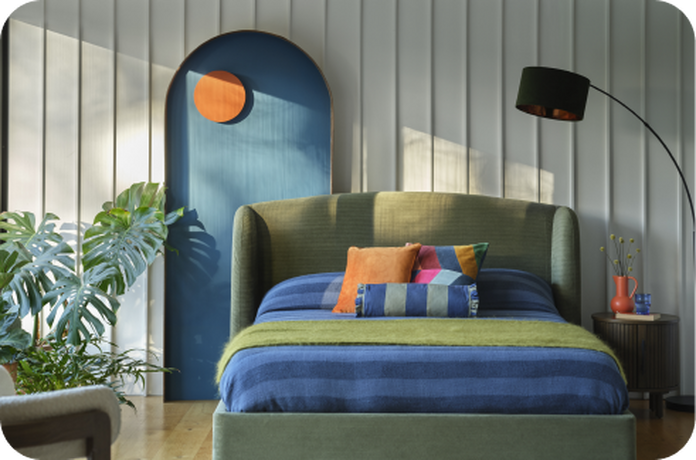 Green divan bed with blue and green bedding against a panelled wall and wooden flooring