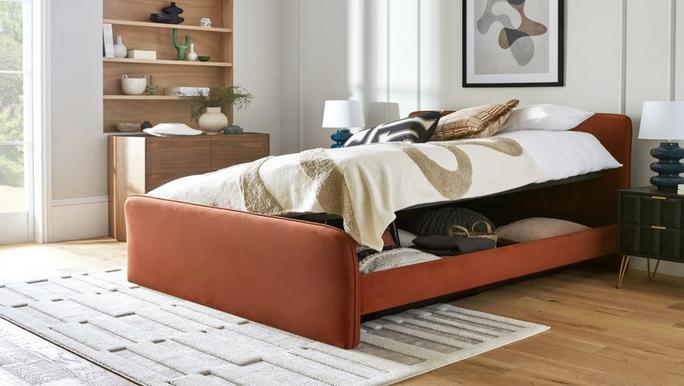 Aro double ottoman bed frame shown with the side opening in rust colour fabric, alongside a black bedside table and neutral rug.