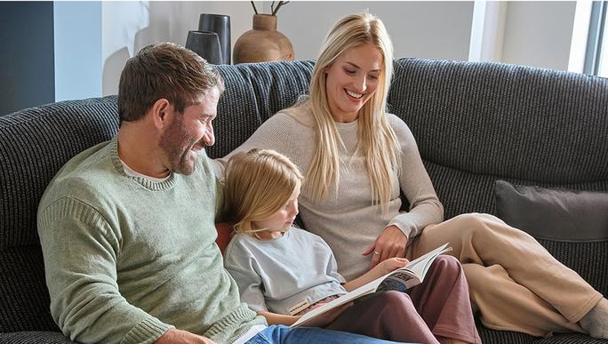 Young family with young child sitting on a sofa laughing at a book
