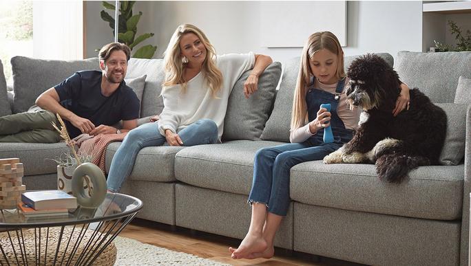young family with small child and dog laughing and playing whilst sat on a sofa 