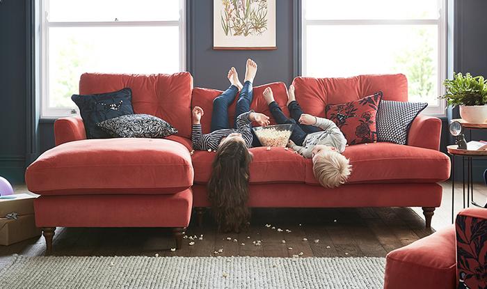 Two young children laying on a red corner sofa making a mess with a bowl of popcorn 
