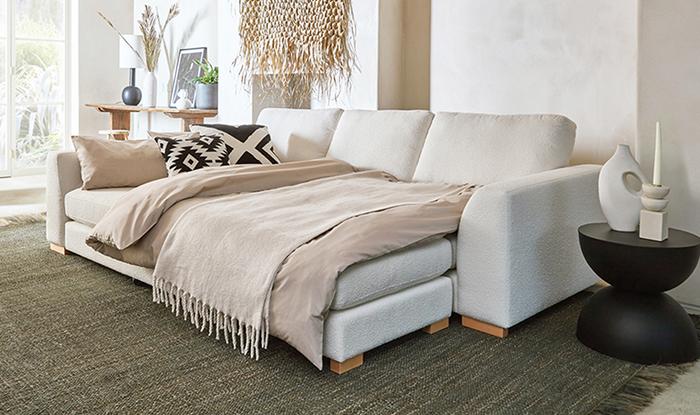 A cosy living room with a white sofa bed, beige bedding and throws with a black side table